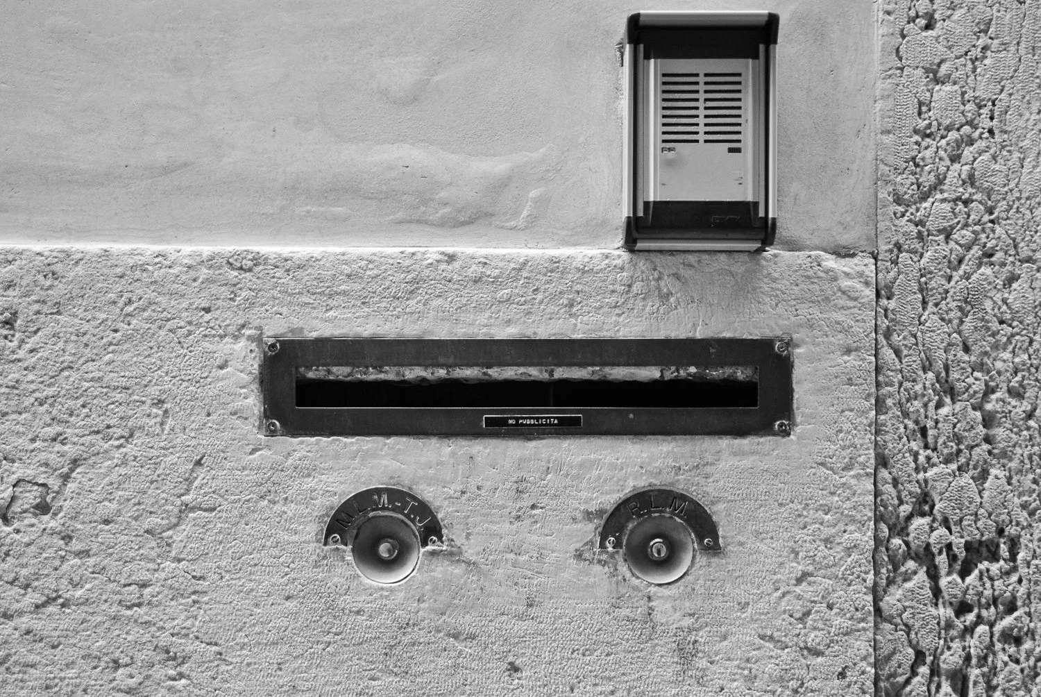 Black and white photo of a building exterior with an mailbox slot, a doorbell, and two circular meters, one labeled P.M.T.U and the other B.C.L.M.