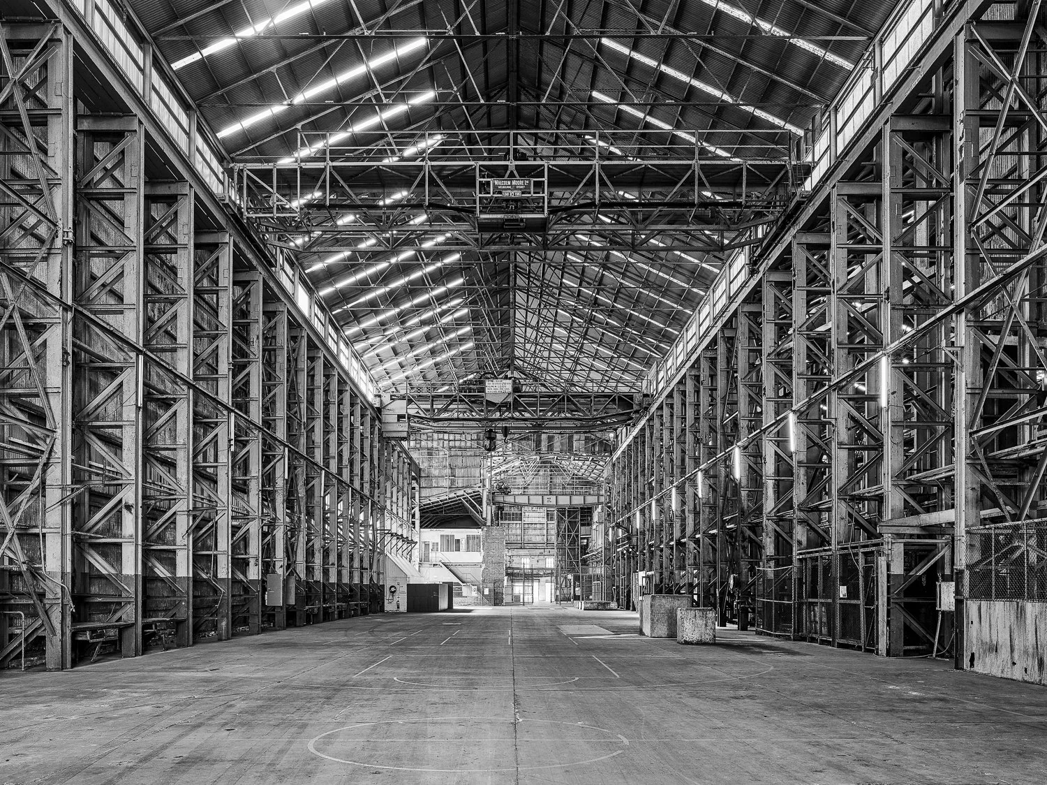 An empty indoor basketball court with a large industrial-style metal structure and lighting, taken in black and white.