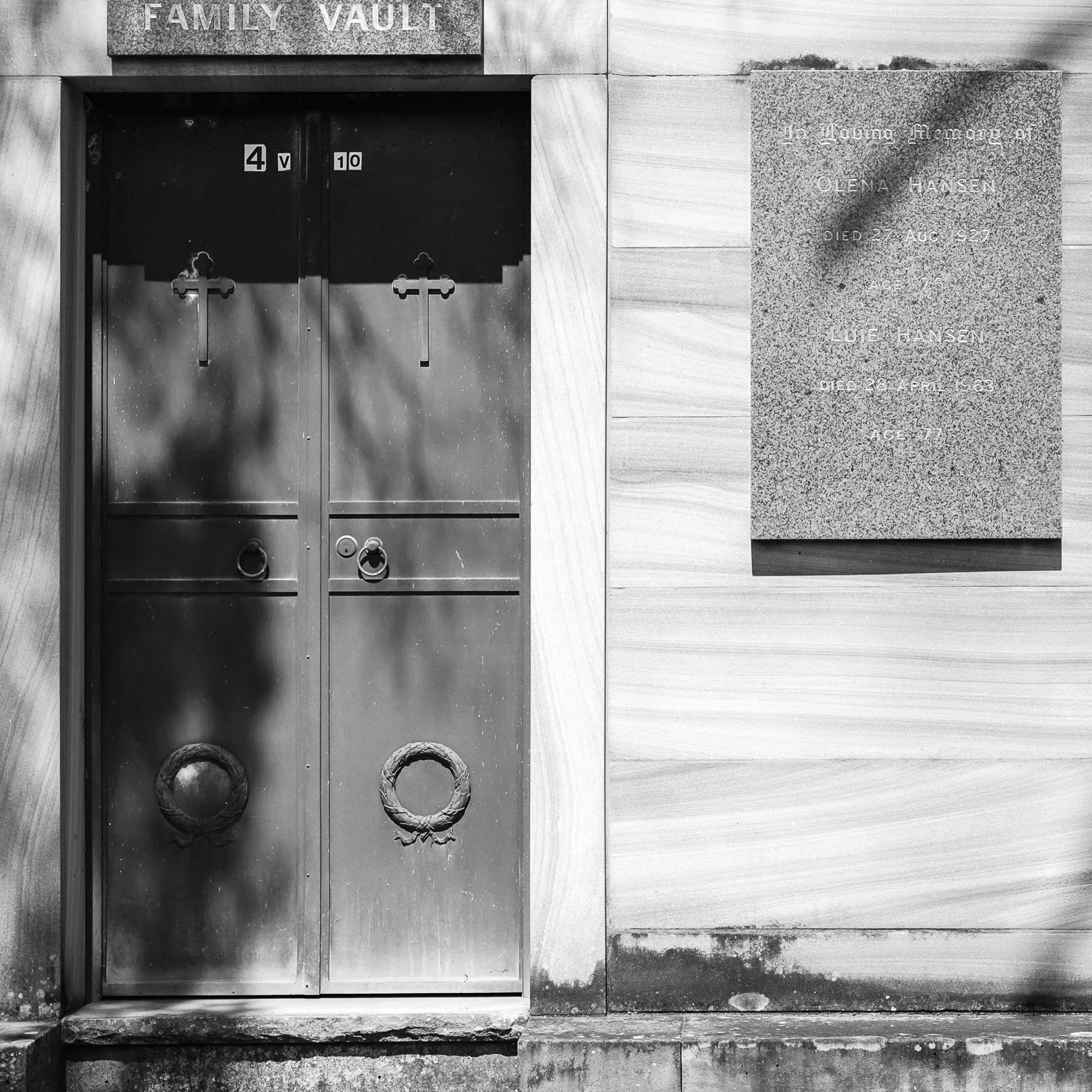 A black and white photo of a metal vault door with two crosses and two circular handles, next to a memorial plaque on a wall.