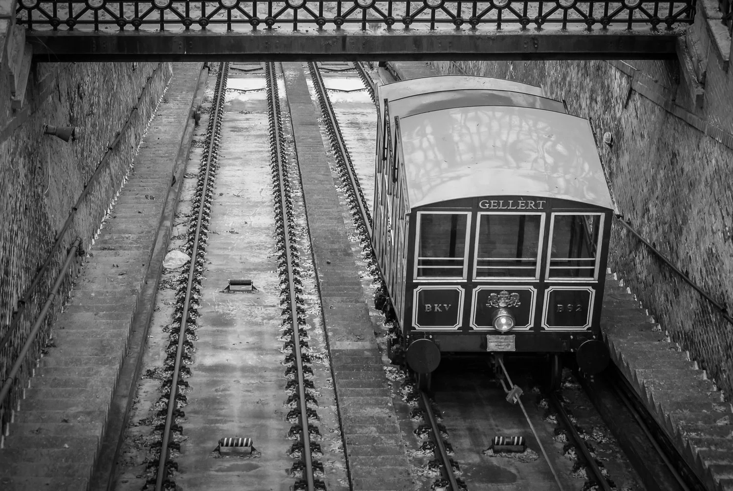A vintage funicular railway car named 'Gellért' at the station, on tracks between brick walls, with a stone pathway on one side and a small slope on the other.