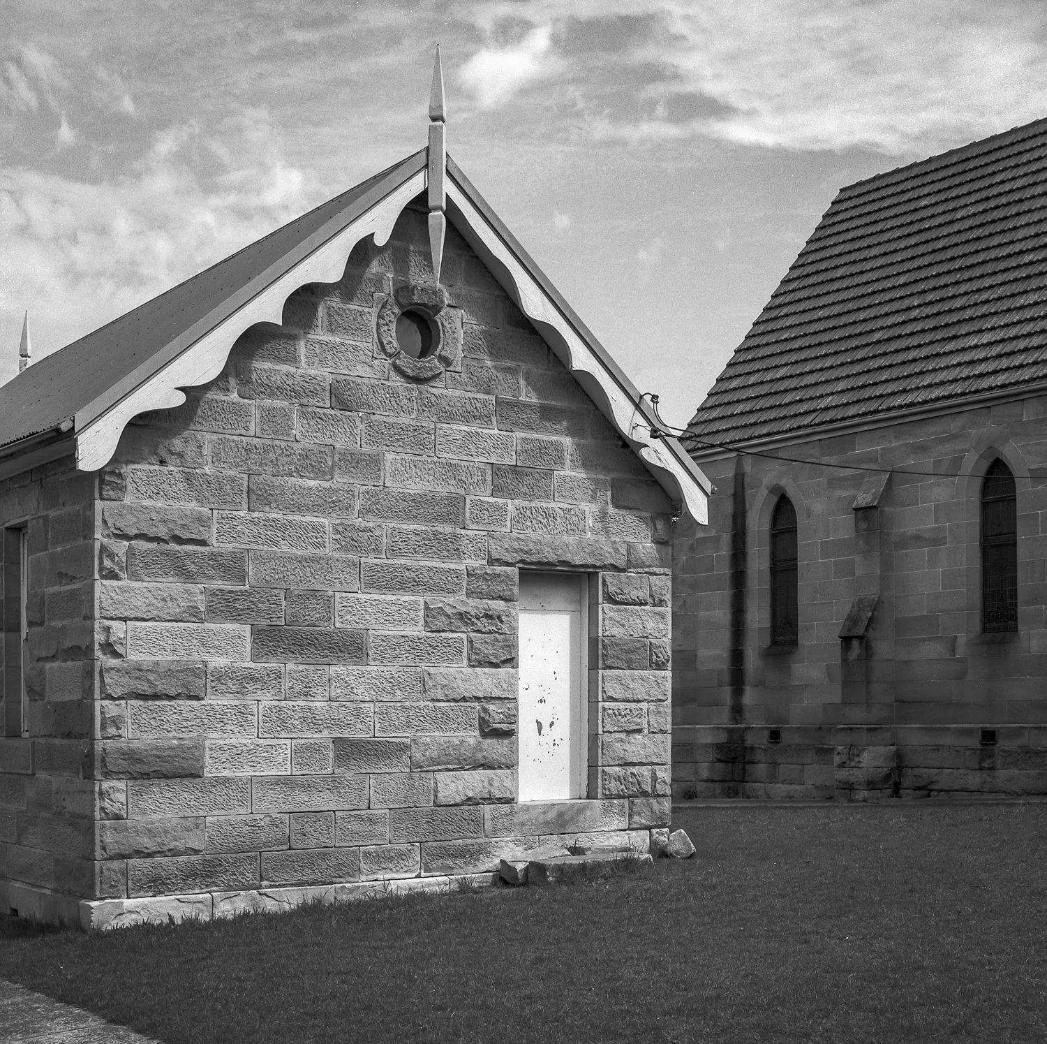 A small stone building with a pitched roof and ornate trim details is next to a larger stone church with arched windows.