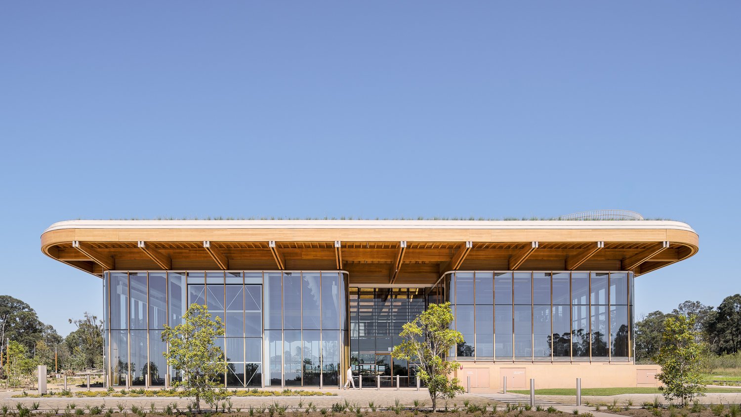Modern glass building with a flat wooden roof, surrounded by trees and a clear blue sky.