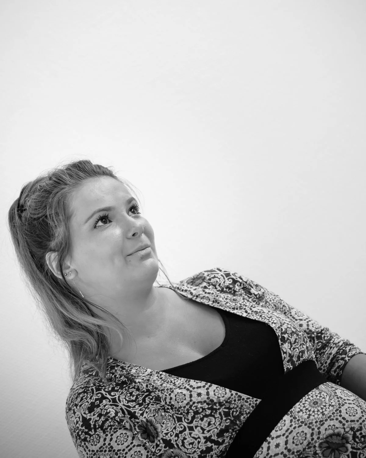 A woman with wavy hair looking upward, wearing a patterned jacket and a black top, against a plain white wall.