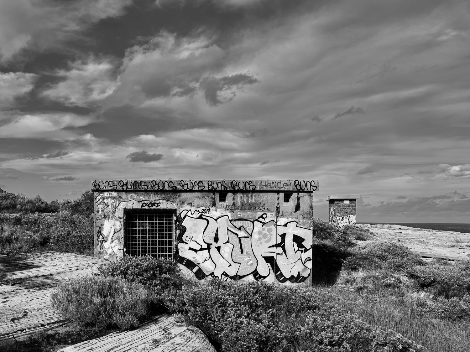 A black and white photo of a small, graffiti-covered concrete building on a beach, with a cloudy sky overhead.