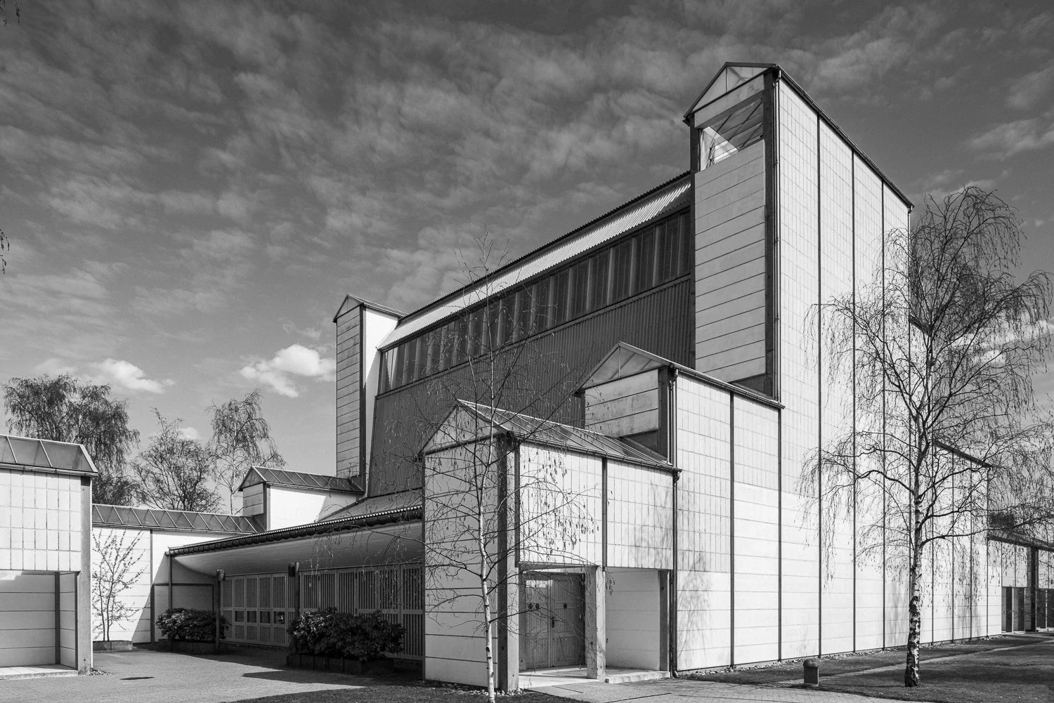 Black and white photo of a modern multi-story building with a combination of flat and sloped roofs and large windows, surrounded by leafless trees.