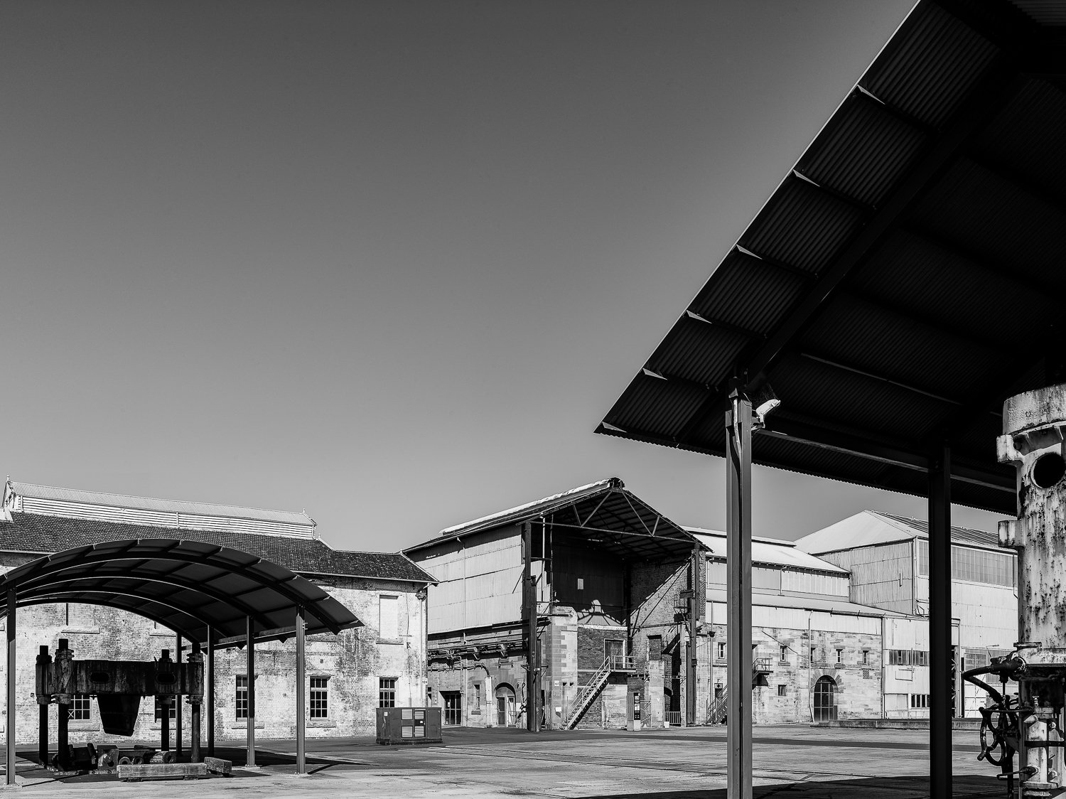 Black and white photo of industrial warehouses and structures, with clear sky and metal roofs.