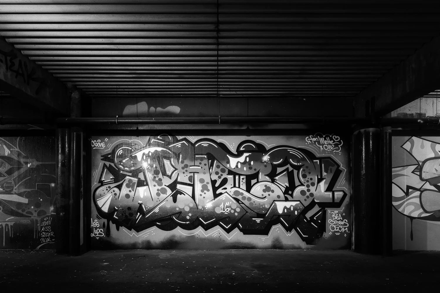 Black and white graffiti art on a wall under an overpass, featuring bold, stylized lettering and various tags.