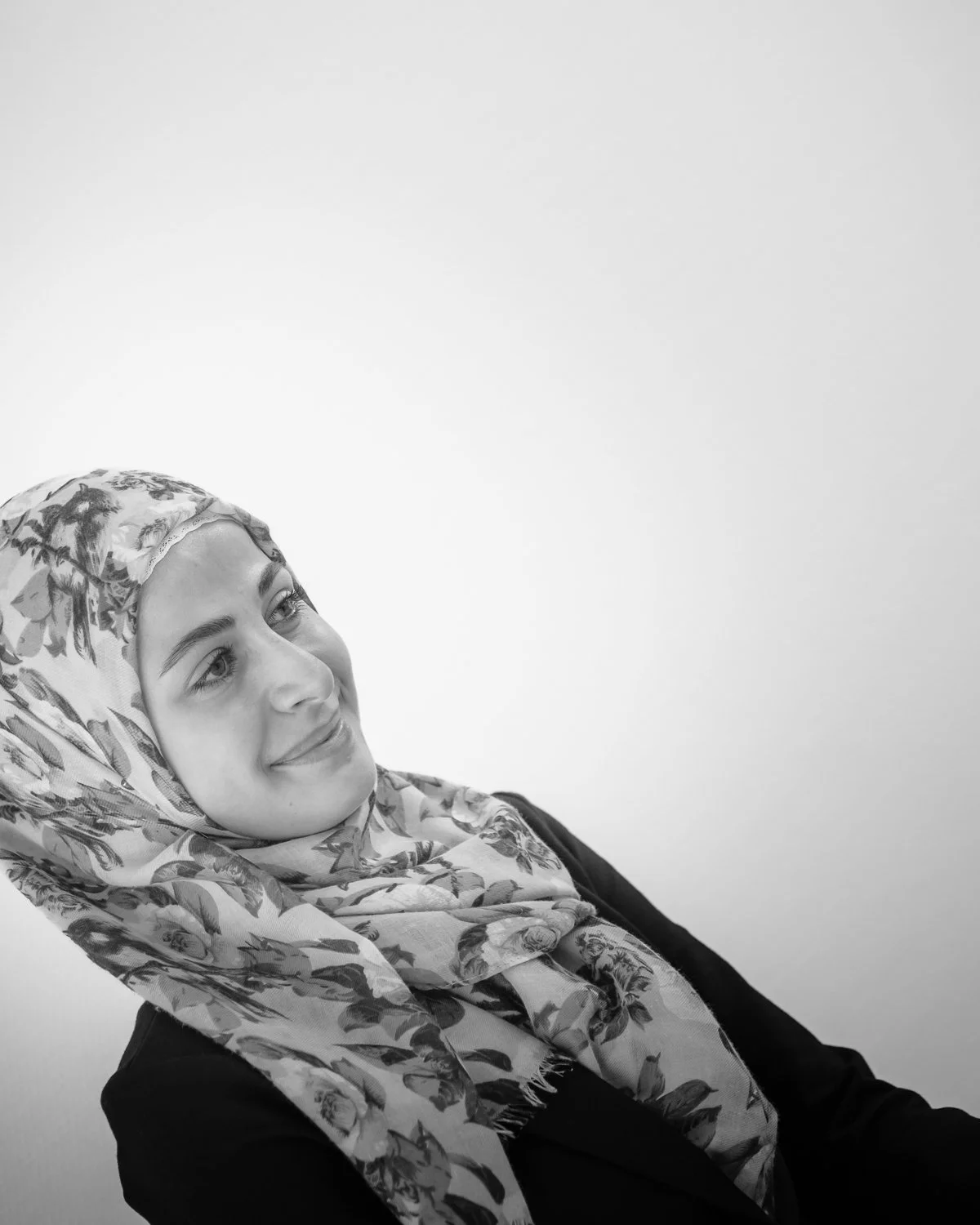 Black and white photo of a woman with a floral patterned headscarf, smiling softly, looking to her right, against a plain light background.
