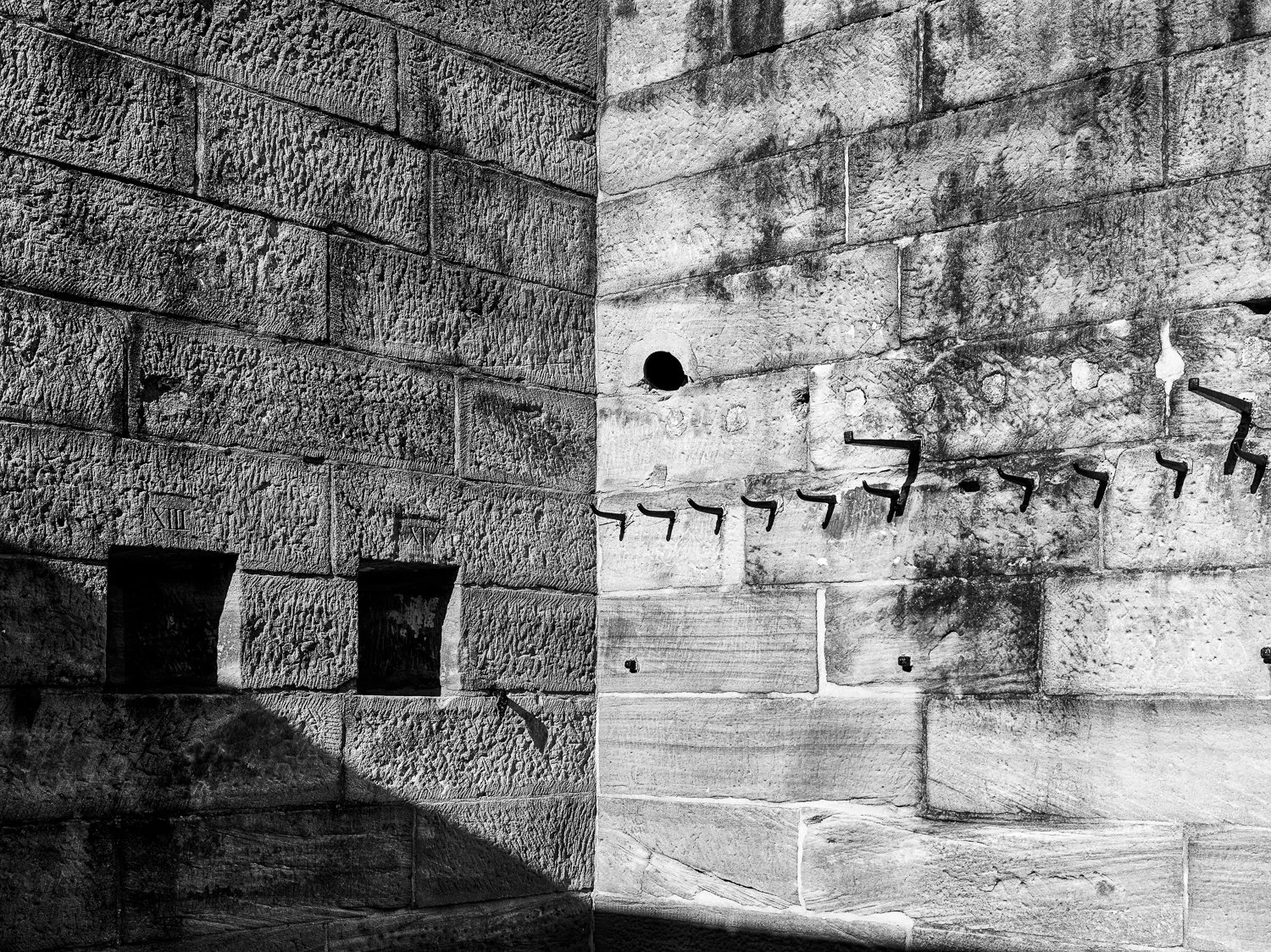 Black and white photo of stone wall with two small windows, a circular hole, and several metal hooks on the right wall, casting shadows.