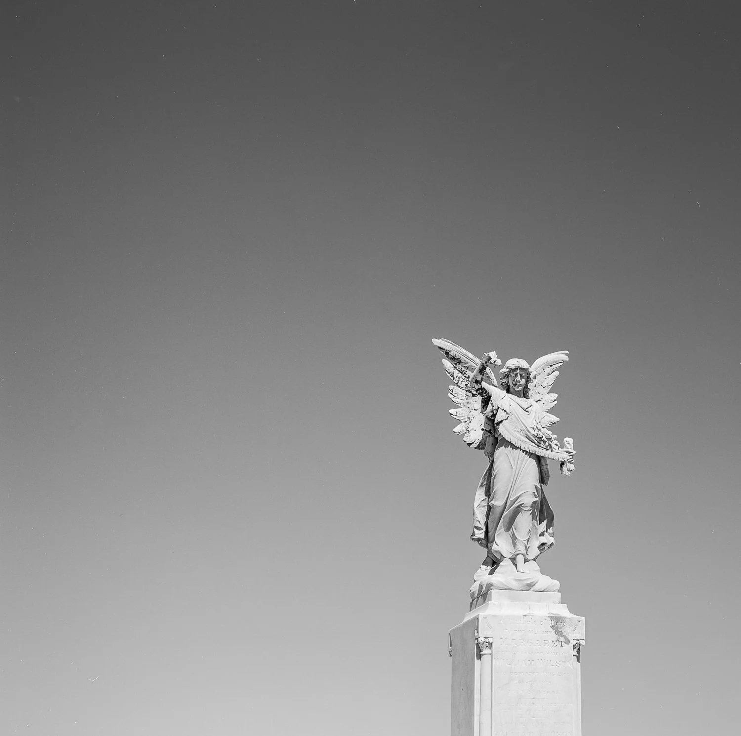 Black and white photograph of a statue of an angel with wings, holding a sword and a scroll, on a pedestal, set against a clear sky with visible stars.
