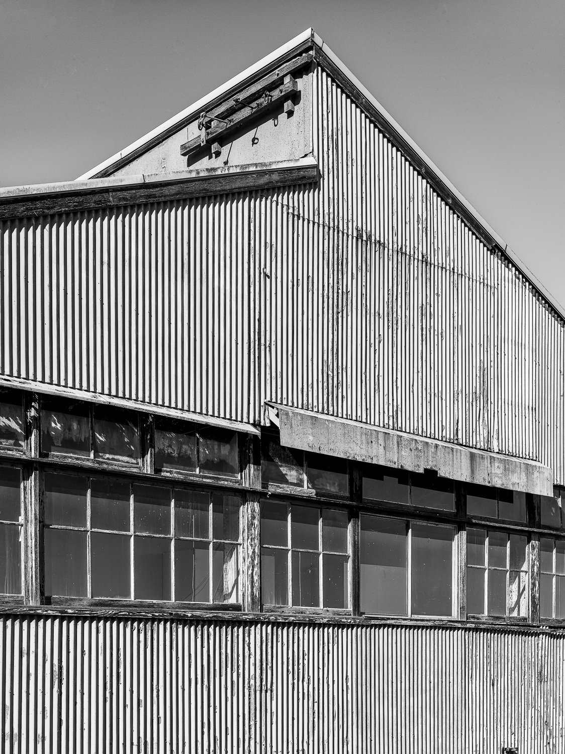Black and white photo of an old, weathered industrial building with corrugated metal siding and large glass windows, showing signs of aging and deterioration.