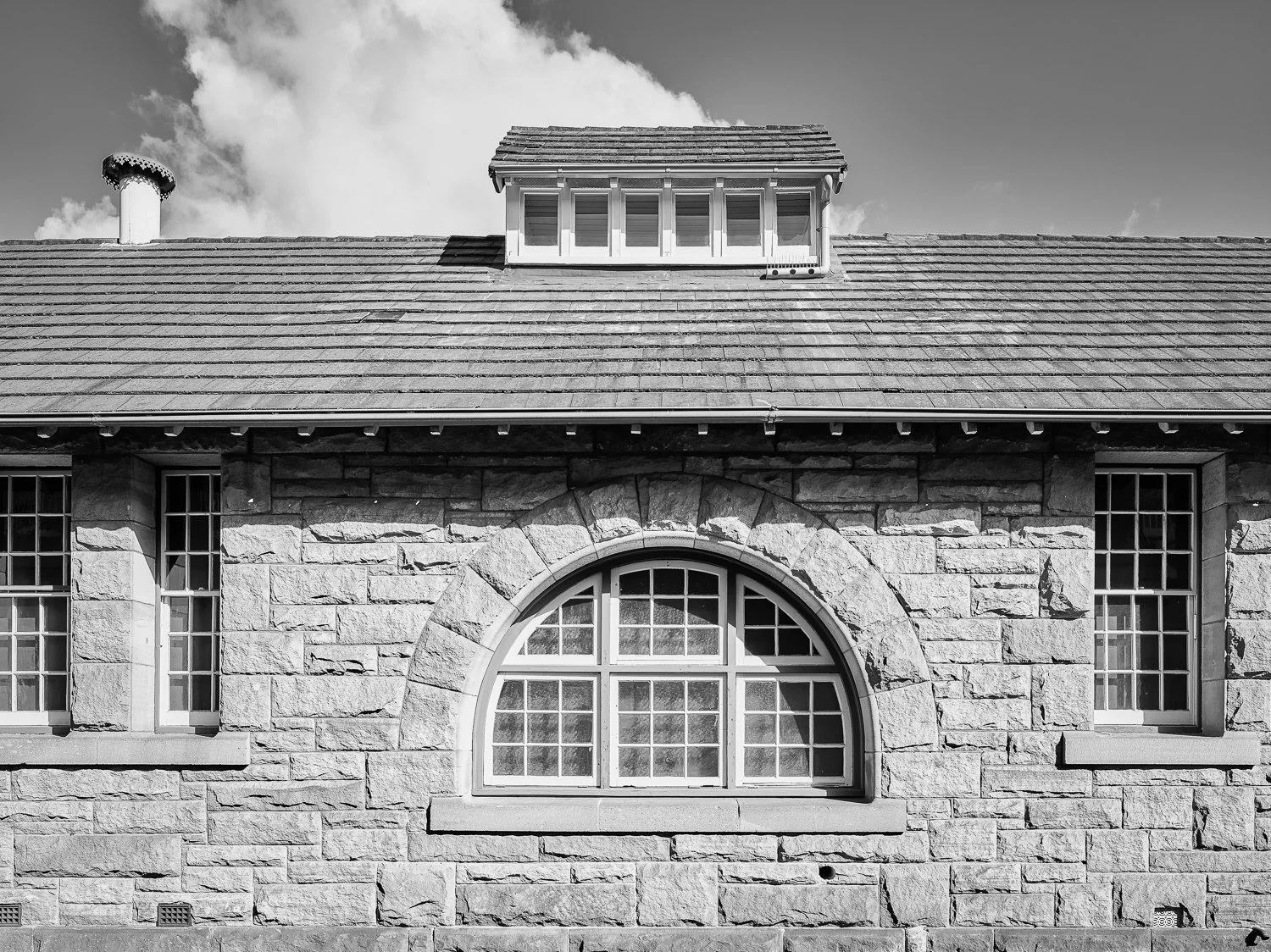 Black and white photo of a stone building with a large arched window and two smaller rectangular windows on either side, a sloped roof with a dormer window on top, and a chimney in the background with a partly cloudy sky.