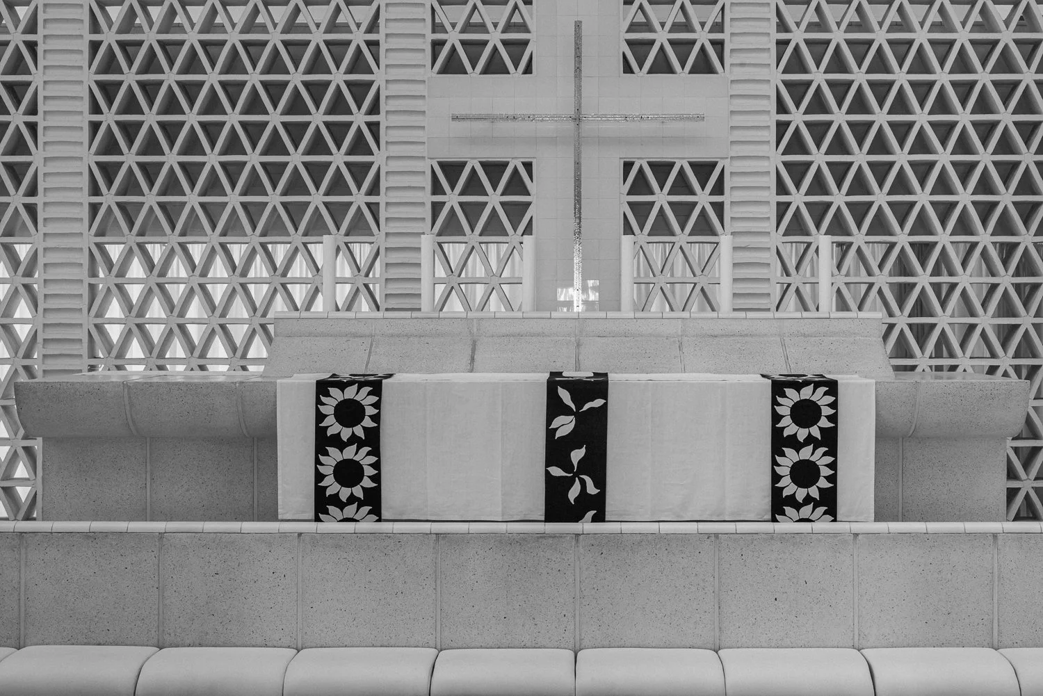 An altar with a cloth featuring sunflower and leaf patterns, set in front of a cross and a wall with a geometric lattice design.