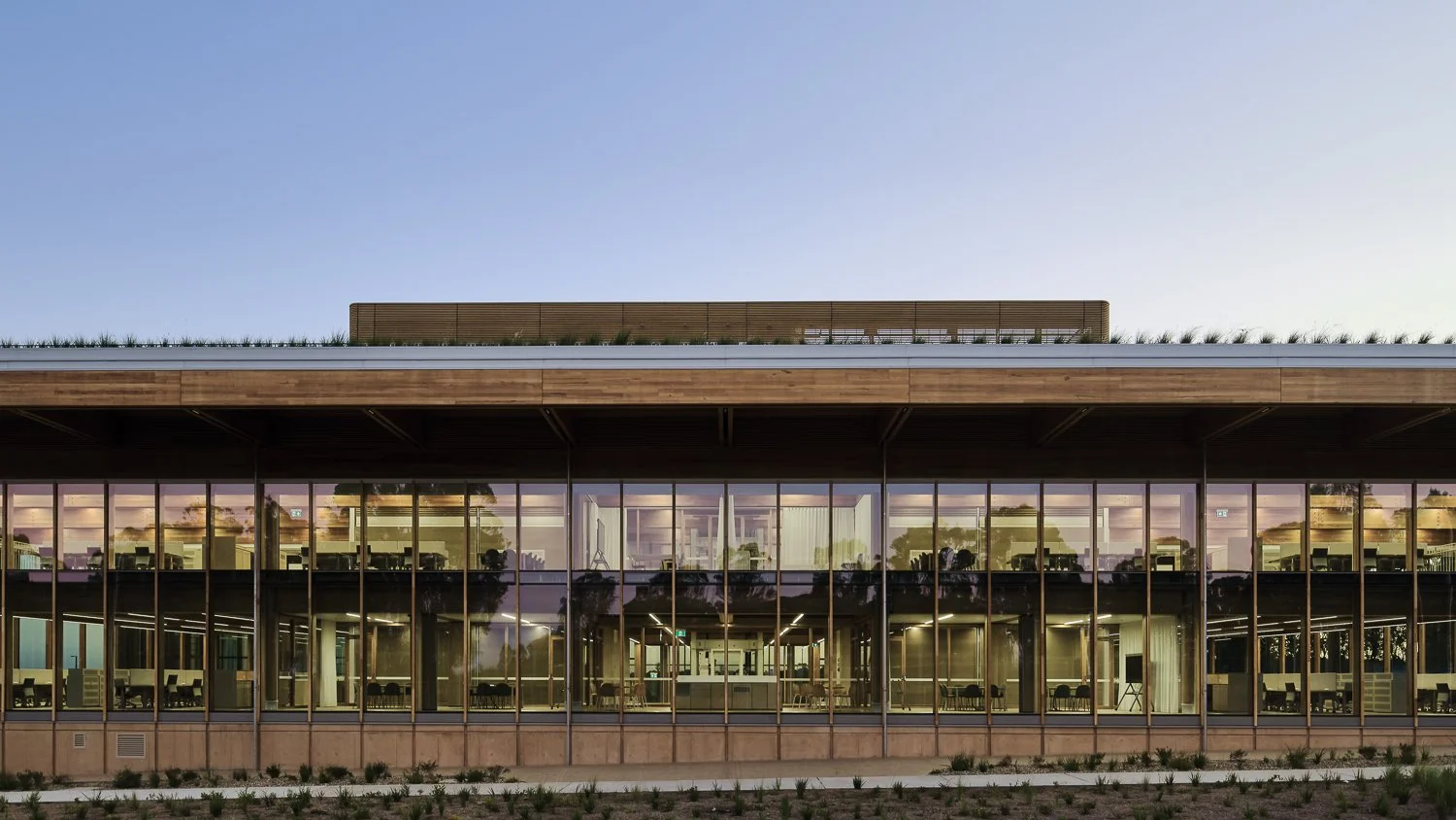 Modern building with large glass windows and a wooden upper facade, illuminated from inside during dusk.