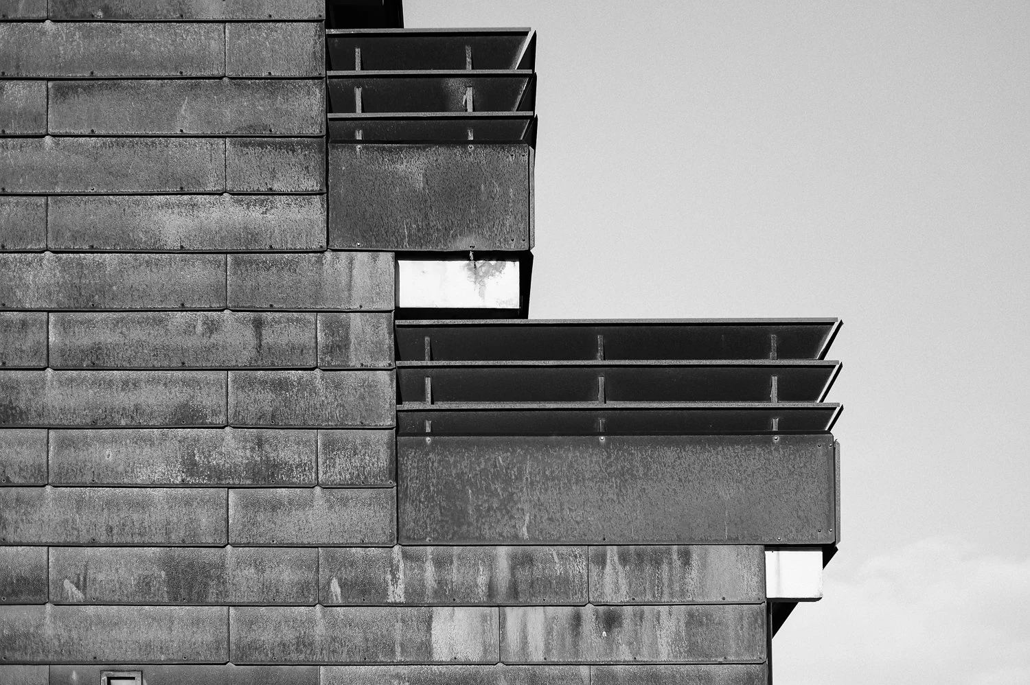 Close-up view of a modern building's exterior showing a textured brick wall on the left and several overlapping, dark-colored metal balcony or architectural panels on the right. The sky is visible in the background.