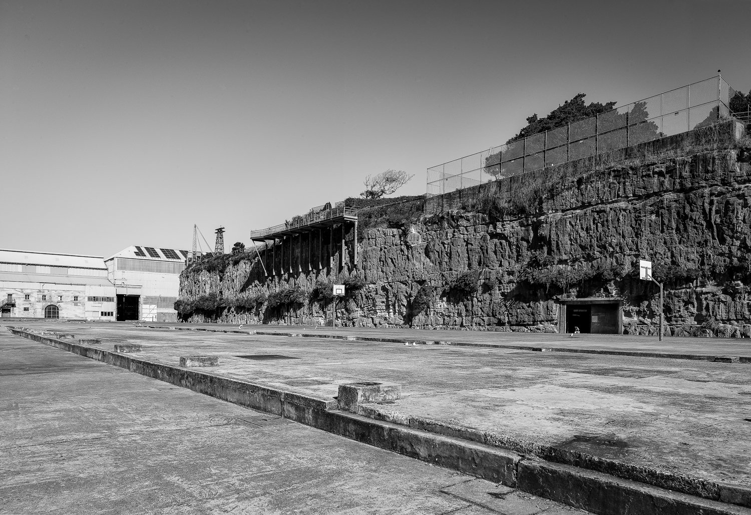 Empty outdoor basketball court with two hoops, surrounded by a rocky cliff with fence on top, in black and white.