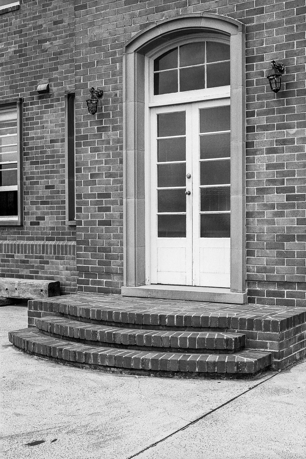 Black and white photo of a brick building entrance with a double door with glass panels, a stone arch, steps, and wall-mounted lanterns.