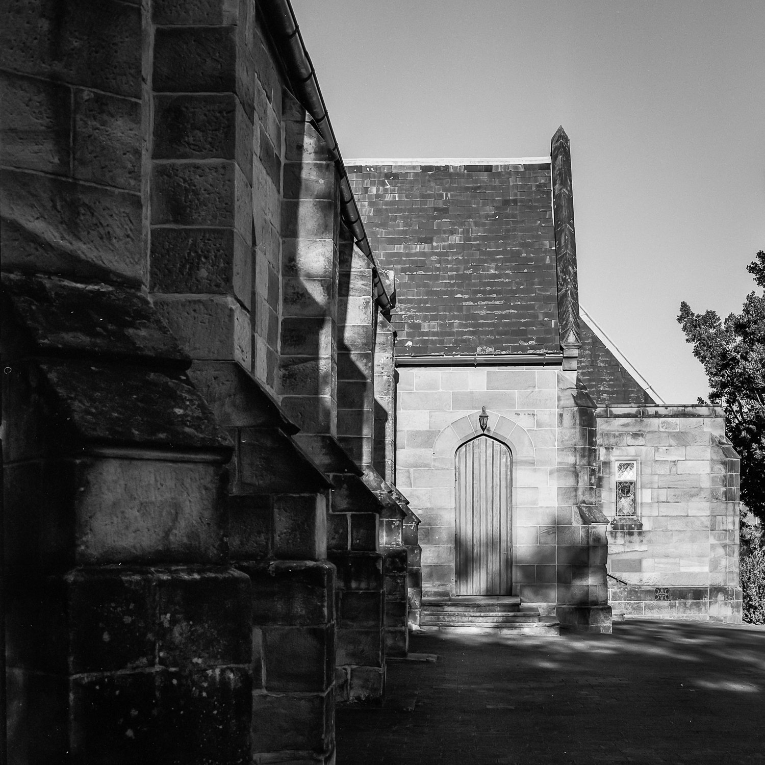 Black and white photo of a stone building with a wooden door, stairs, and small window, partially shaded by trees.