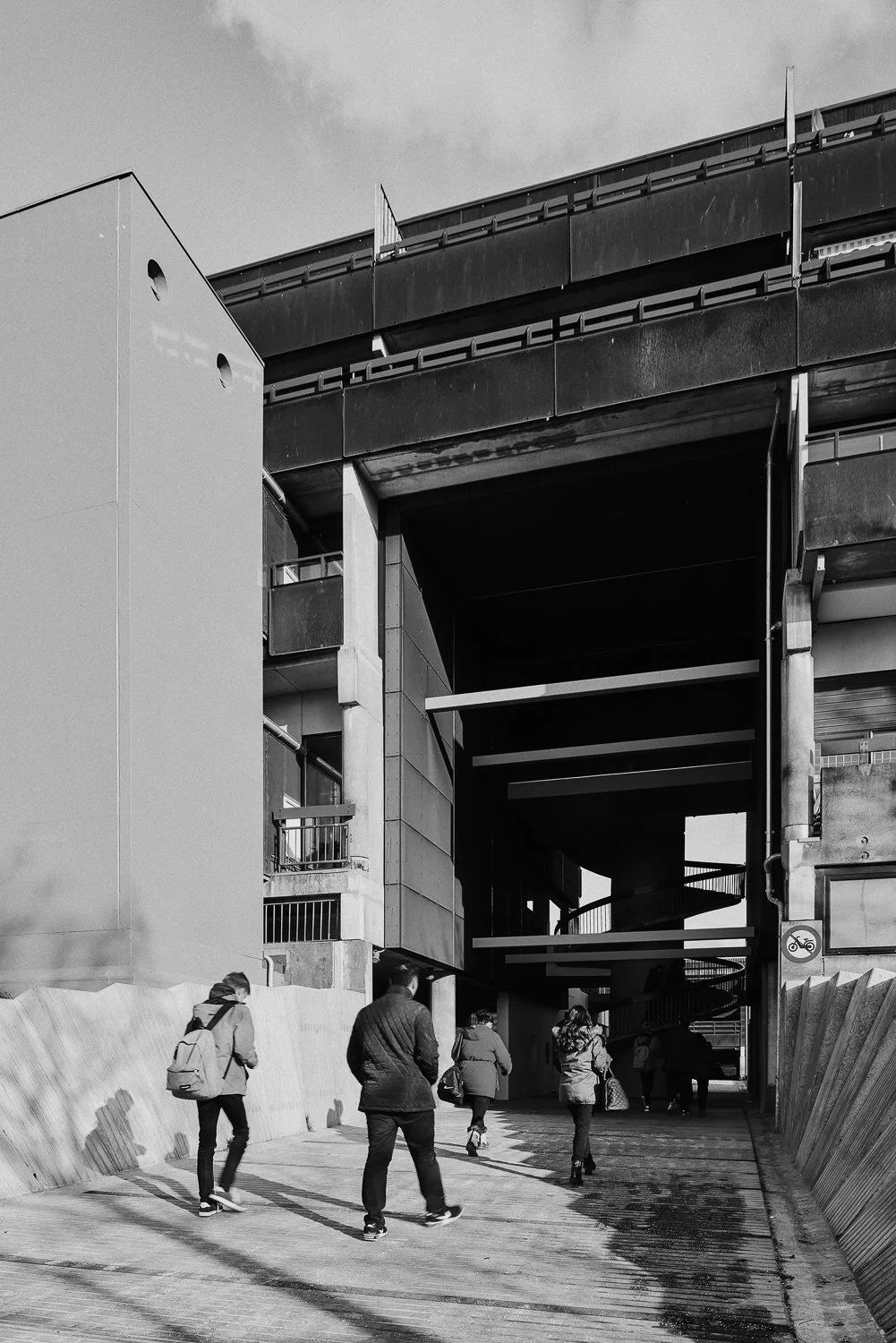 Black and white photo of pedestrians walking under a modern building with multiple levels and staircases.