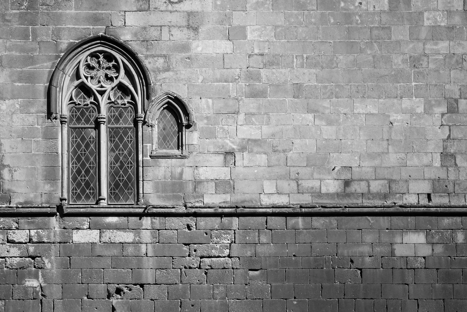 Black and white photo of a stone brick wall with two Gothic-style arched windows, one larger with intricate tracery and diamond leaded glass, and a smaller window beside it.