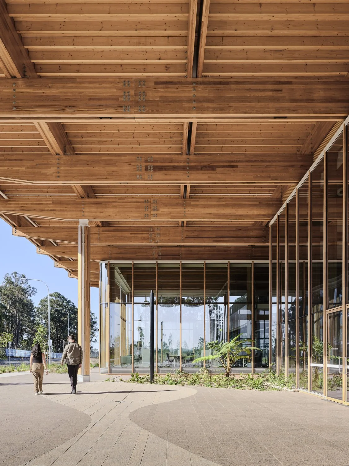 Modern building with a wooden overhang, glass walls, and small plants outside. Two people walking toward the building.