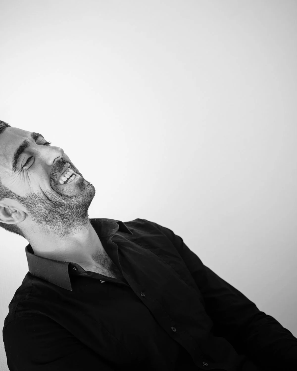 Black and white photo of a man laughing with eyes closed, head tilted back, wearing a dark button-up shirt, against a plain light background.