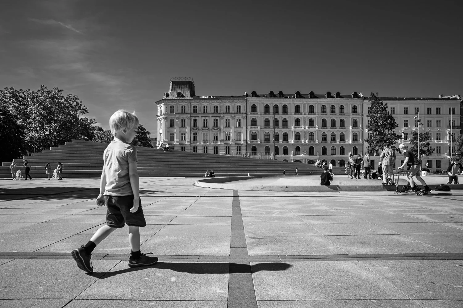 A black and white photo of a young boy walking on a paved square with a large staircase and a historic building in the background. There are groups of people, some sitting and some standing, around the area, and two kids riding scooters.