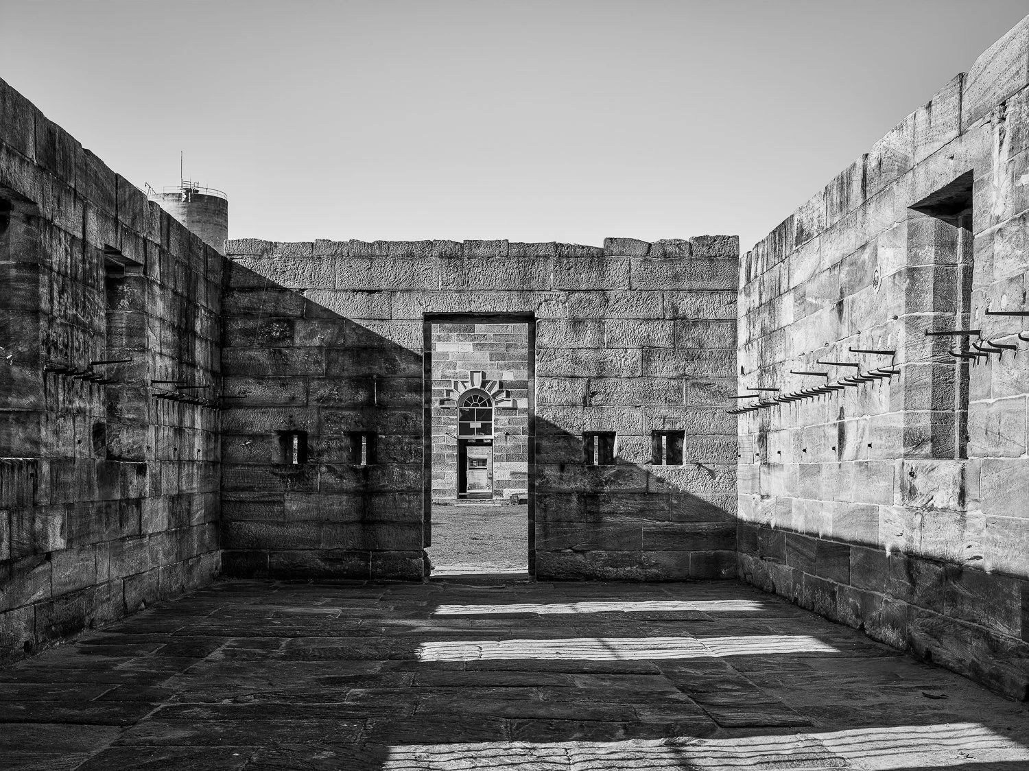 Black and white photo of an old stone fortification wall with a central arched doorway and small rectangular windows. The wall has a pattern of bricks and shadows cast on the ground.