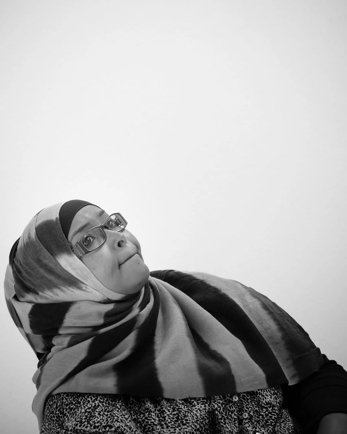 A woman with glasses wearing a hijab, looking thoughtfully to the side, black-and-white photo.