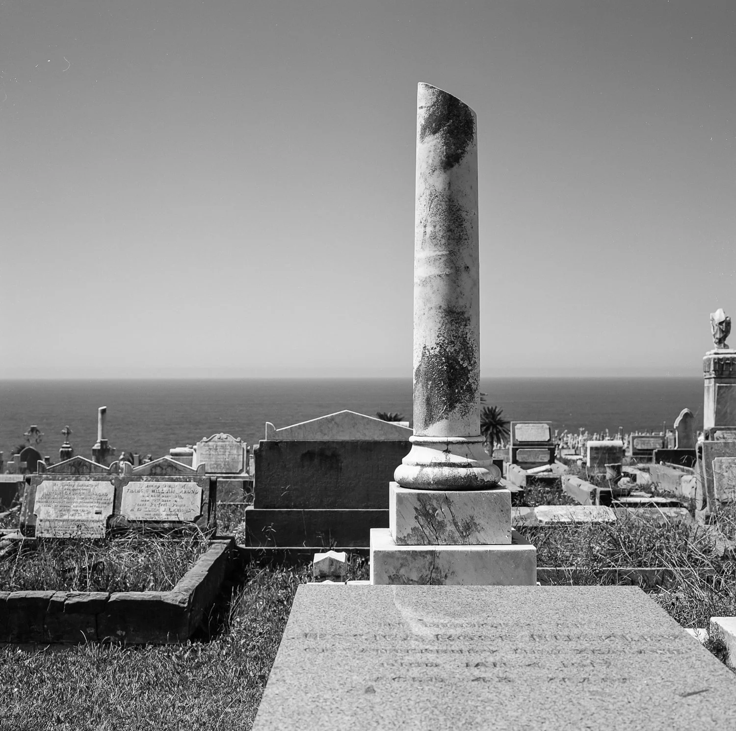Black and white photograph of an old cemetery with a tall, weathered marble column monument in the foreground, and the ocean with a clear sky in the background.