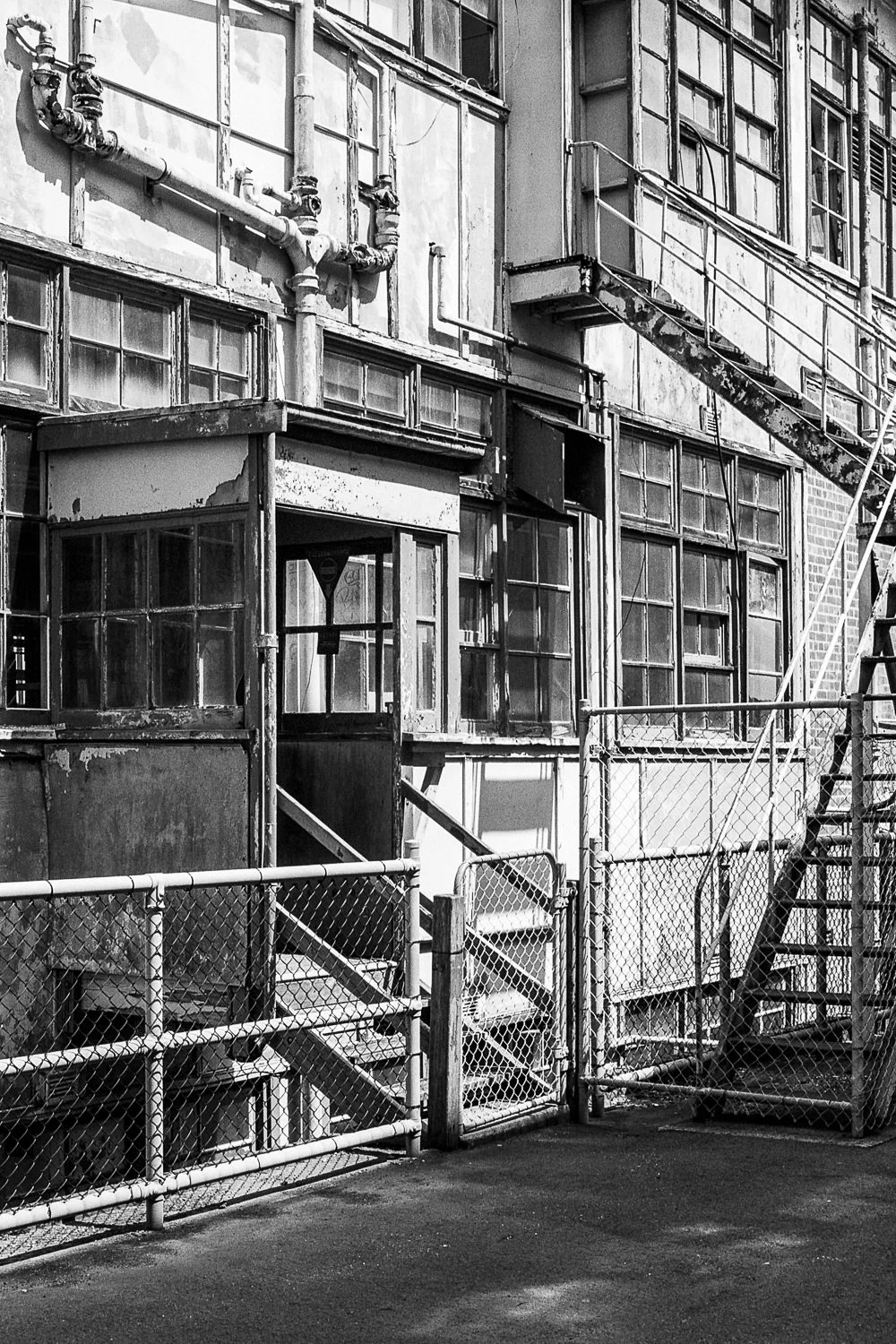 Black and white photo of an old, weathered brick building with numerous large, multi-pane windows, a fire escape ladder, pipes, and a chain-link fence in the foreground.