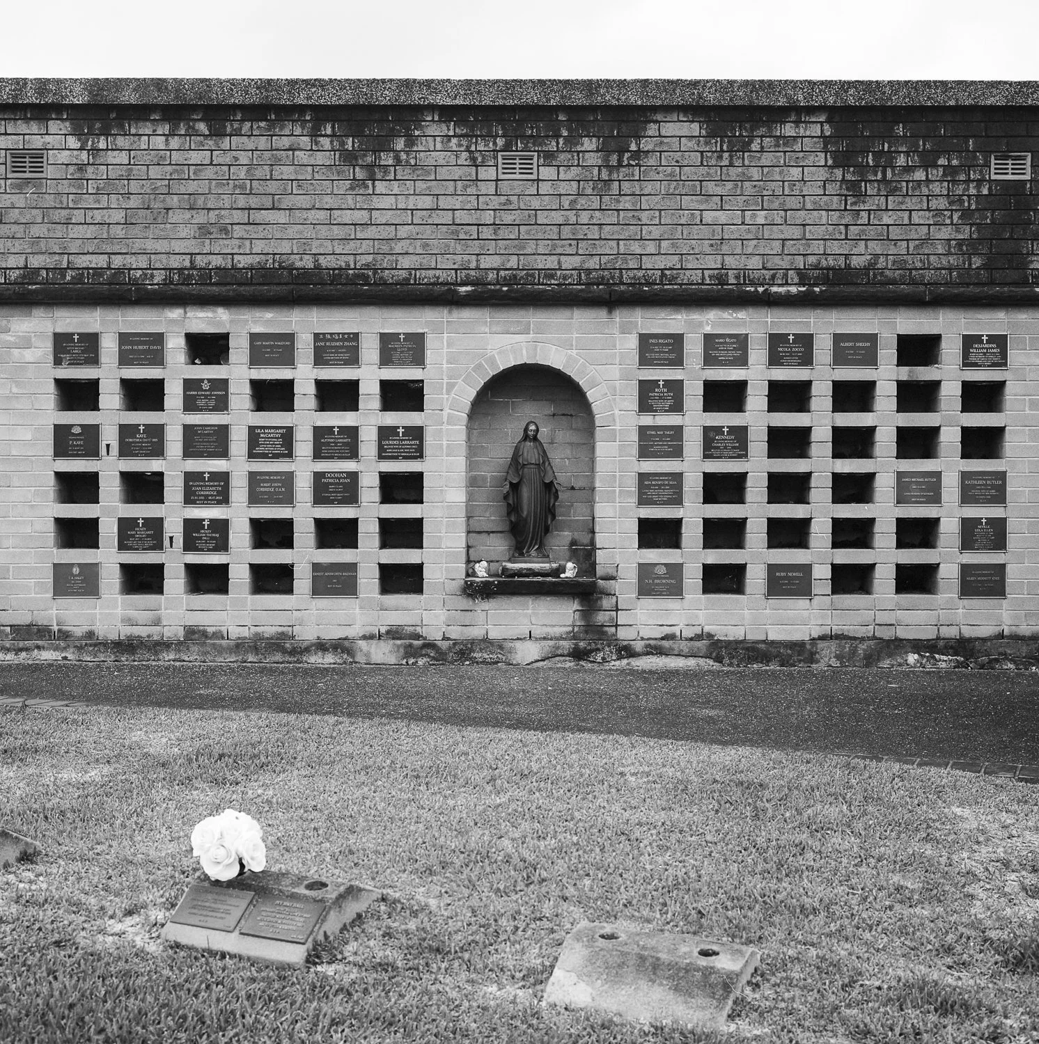 Black and white photo of a memorial wall with plaques and a central statue of the Virgin Mary inside a small niche. There are flowers on a plaque in the grass in front of the wall.
