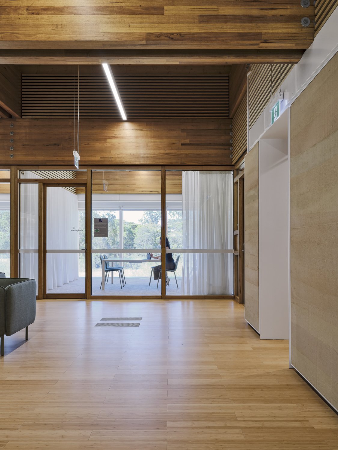 Modern interior space with wooden ceiling and floors, featuring glass partitions, a minimalistic seating area, and a person sitting at a table in the background.