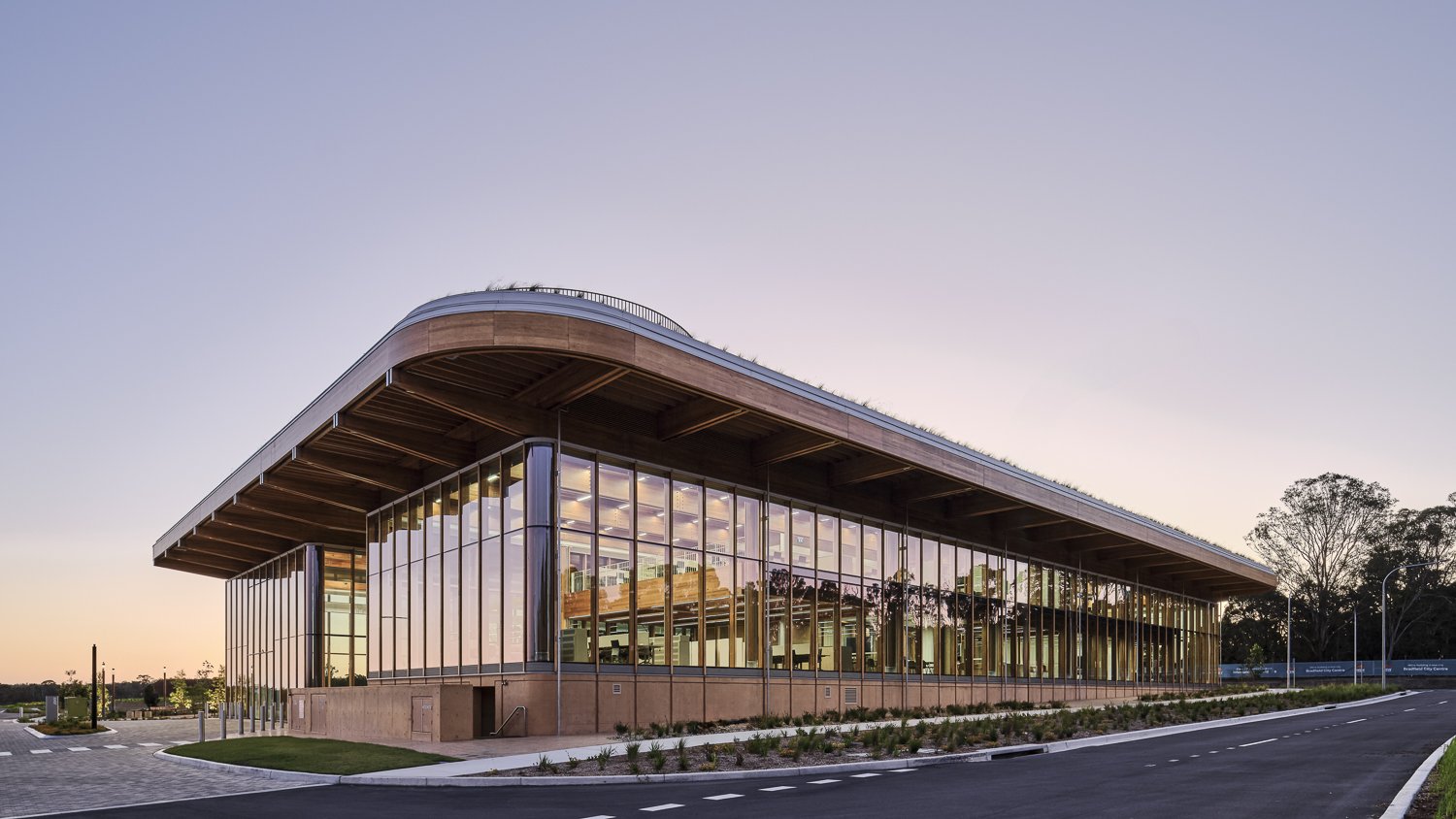 Modern glass building with extensive window panels and a curved roof, surrounded by a paved parking area and landscaped greenery, at dusk.