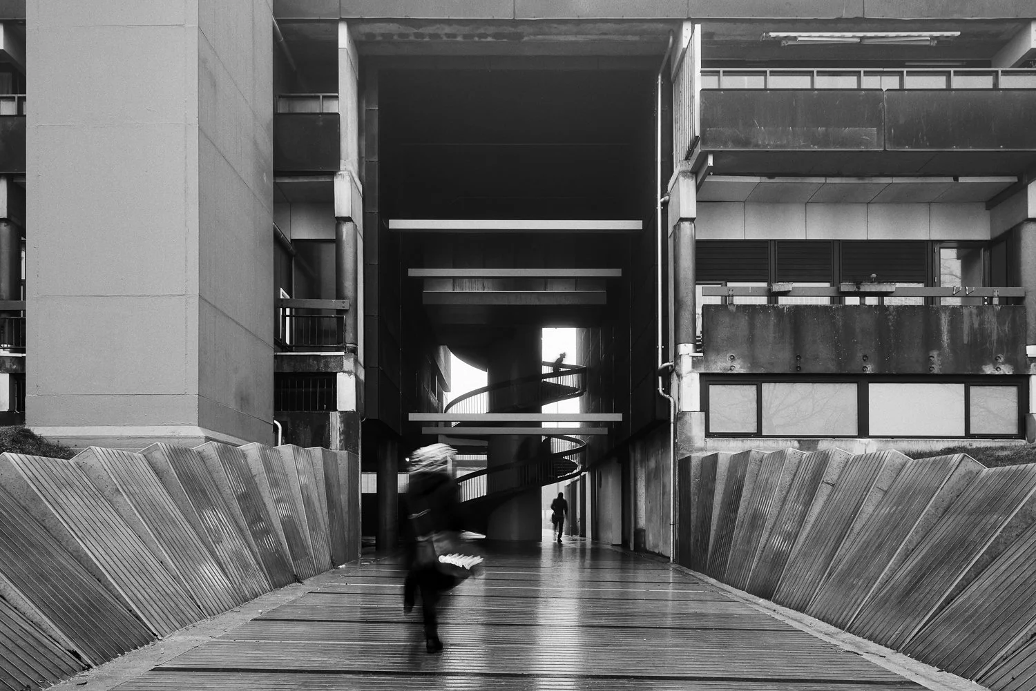 Black and white photo of a modern urban building with an outdoor staircase, a corridor, and pedestrians walking, one blurred in motion.