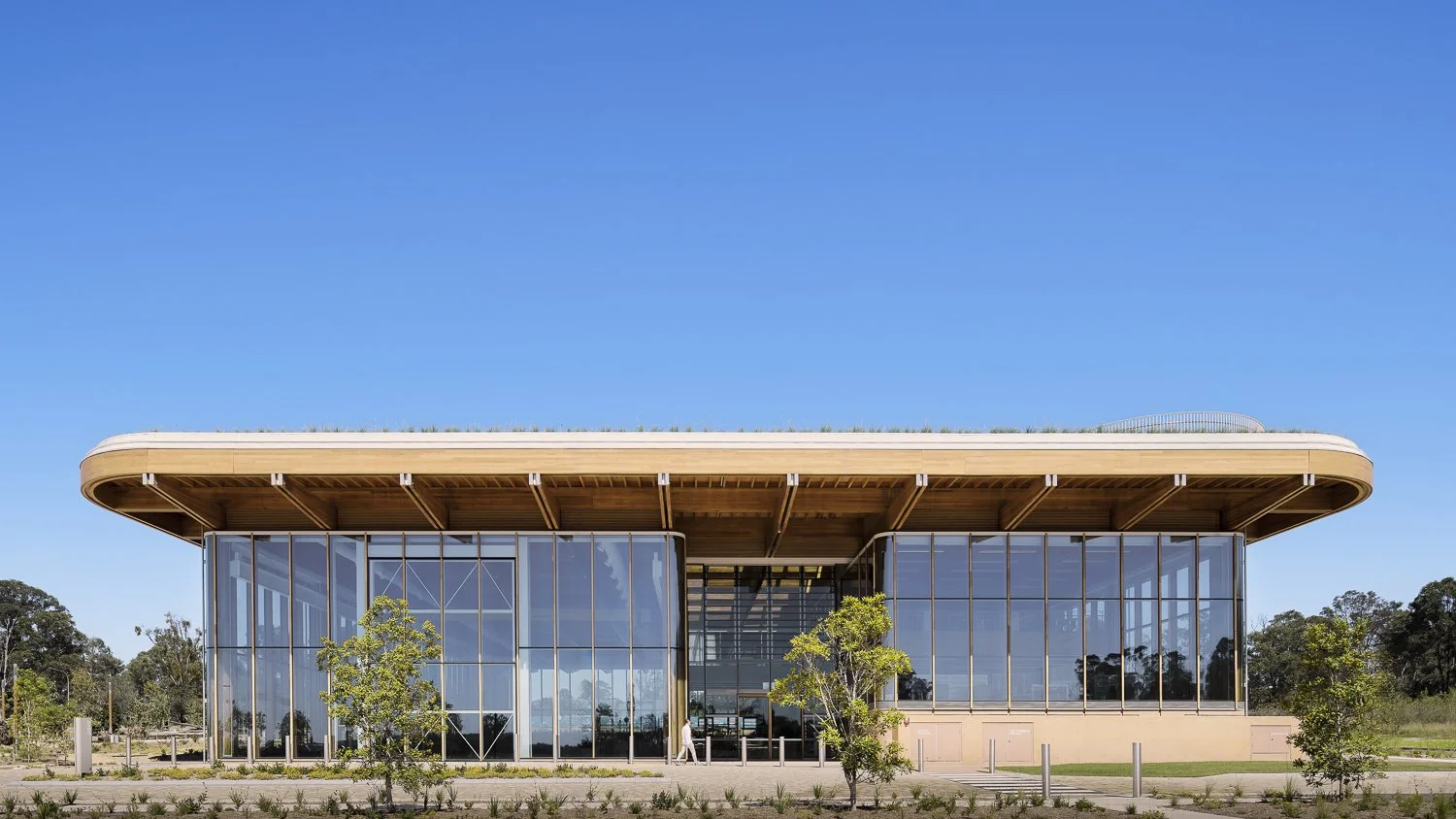Modern building with large glass windows, a curved wooden roof, and surrounding greenery under a clear blue sky.