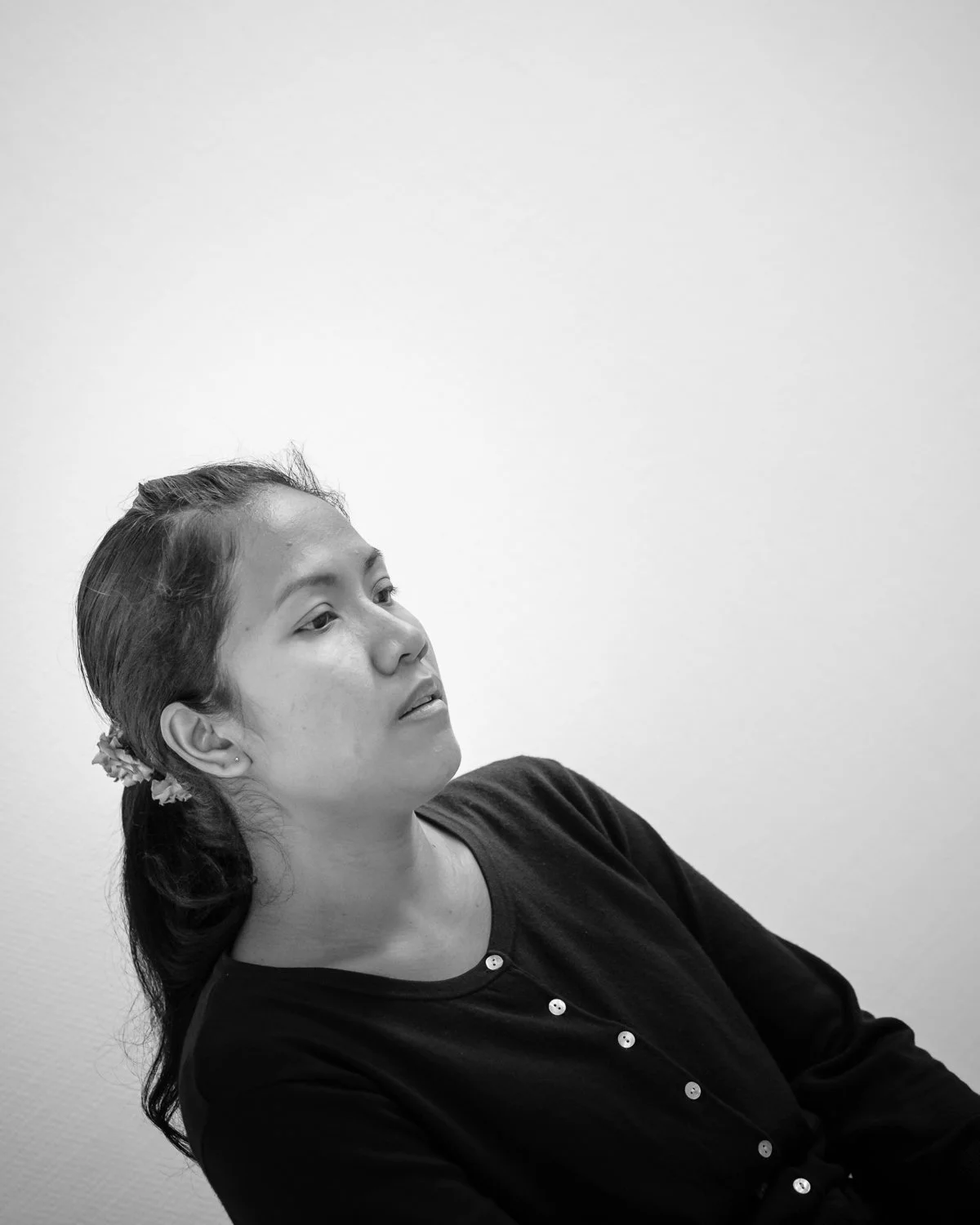 Black and white photo of a woman with long hair tied back, wearing a dark top, sitting against a plain wall, looking to the left with a thoughtful expression.