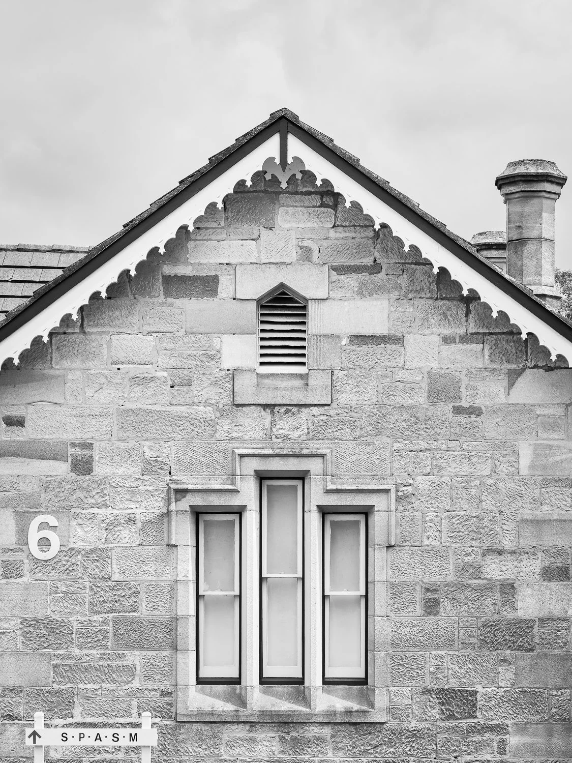 Black and white photo of a house with stone exterior, triangular roof, chimney, and three tall windows on the front wall. Official-looking sign in the bottom left corner with the text "S P A S M" and an arrow pointing up.