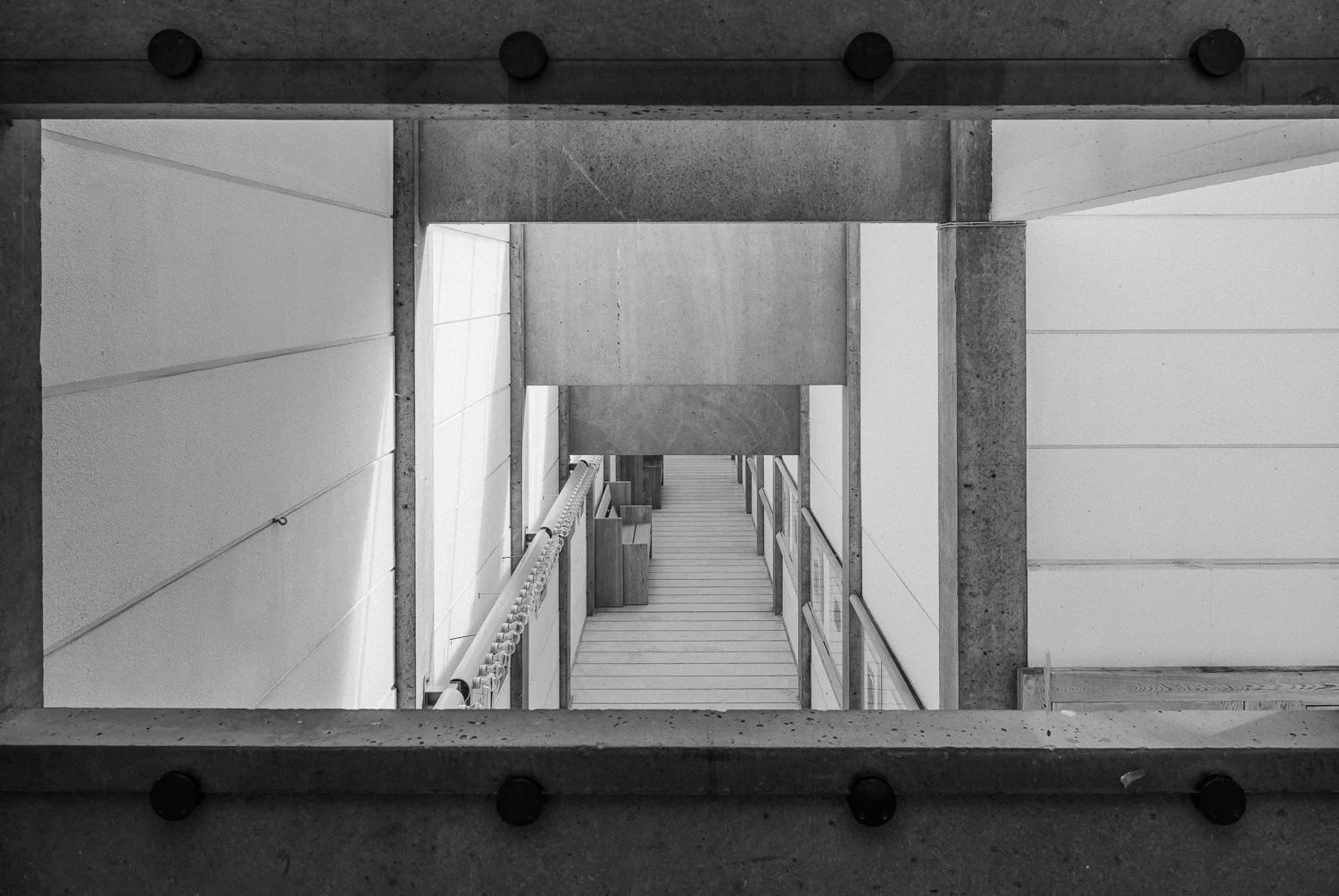 View looking down at a modern stairwell with concrete and white walls, featuring a wooden staircase with metal railings.