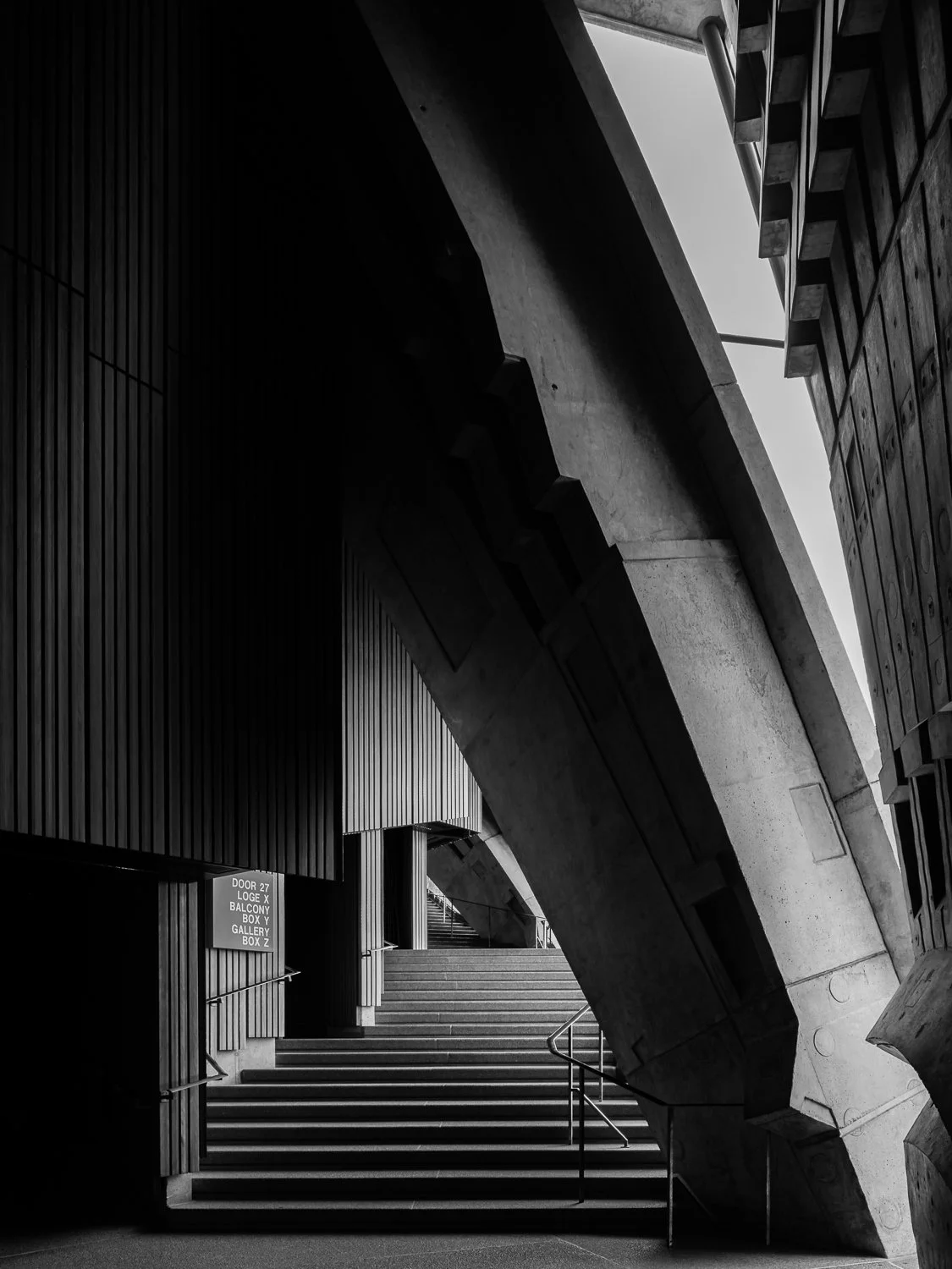 Black and white photo of an modern architectural interior with stairs and concrete walls, featuring a geometric design and a sign indicating different sections.