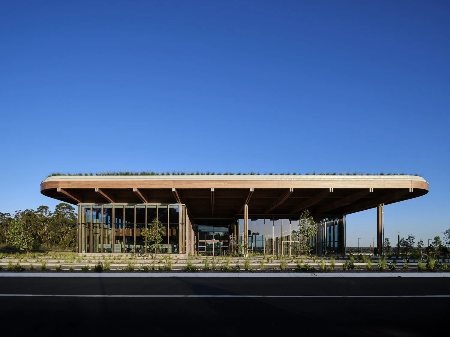 Modern building with large glass windows, curved roof with greenery on top, surrounded by landscaped plants, under a clear blue sky.