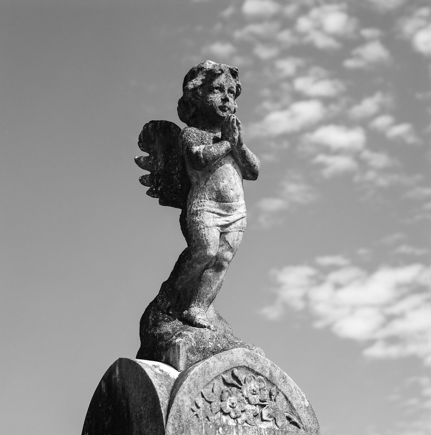 A weathered stone statue of a child angel with wings, standing with hands pressed together in prayer, atop a circular base with floral carvings, against a cloudy sky.