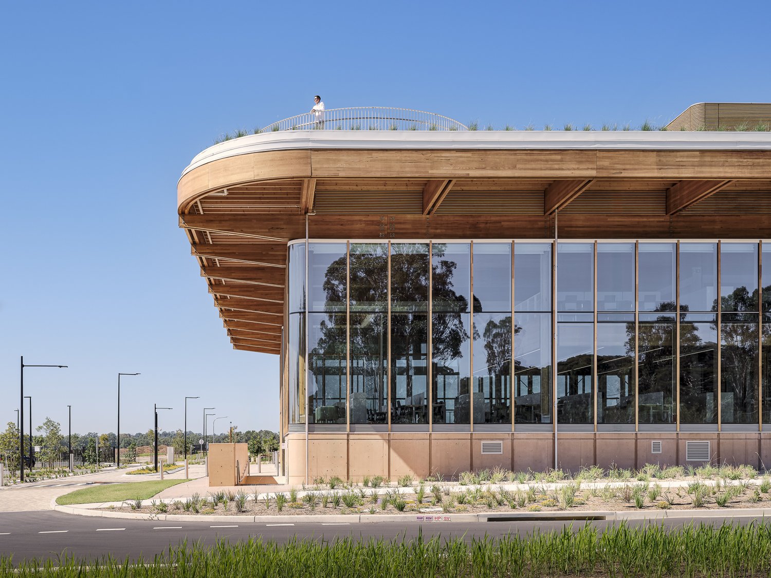 Modern building with large glass windows and wooden accents, featuring a person standing on a rooftop terrace. The setting is urban with a clear blue sky and nearby greenery.
