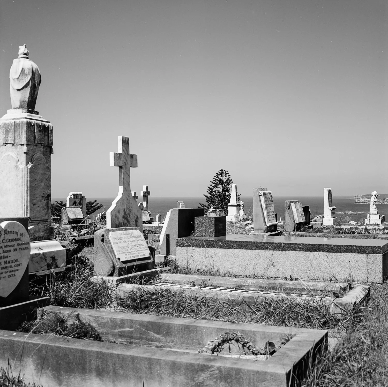 Graveyard with various headstones and crosses, some with engraved inscriptions, overlooking the ocean with a tree and clear sky in the background.