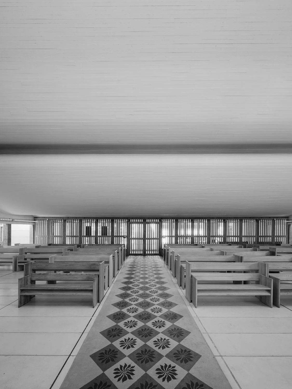 Black and white photo of the interior of a church or chapel with wooden pews, a decorative tiled aisle, and wooden slatted walls.