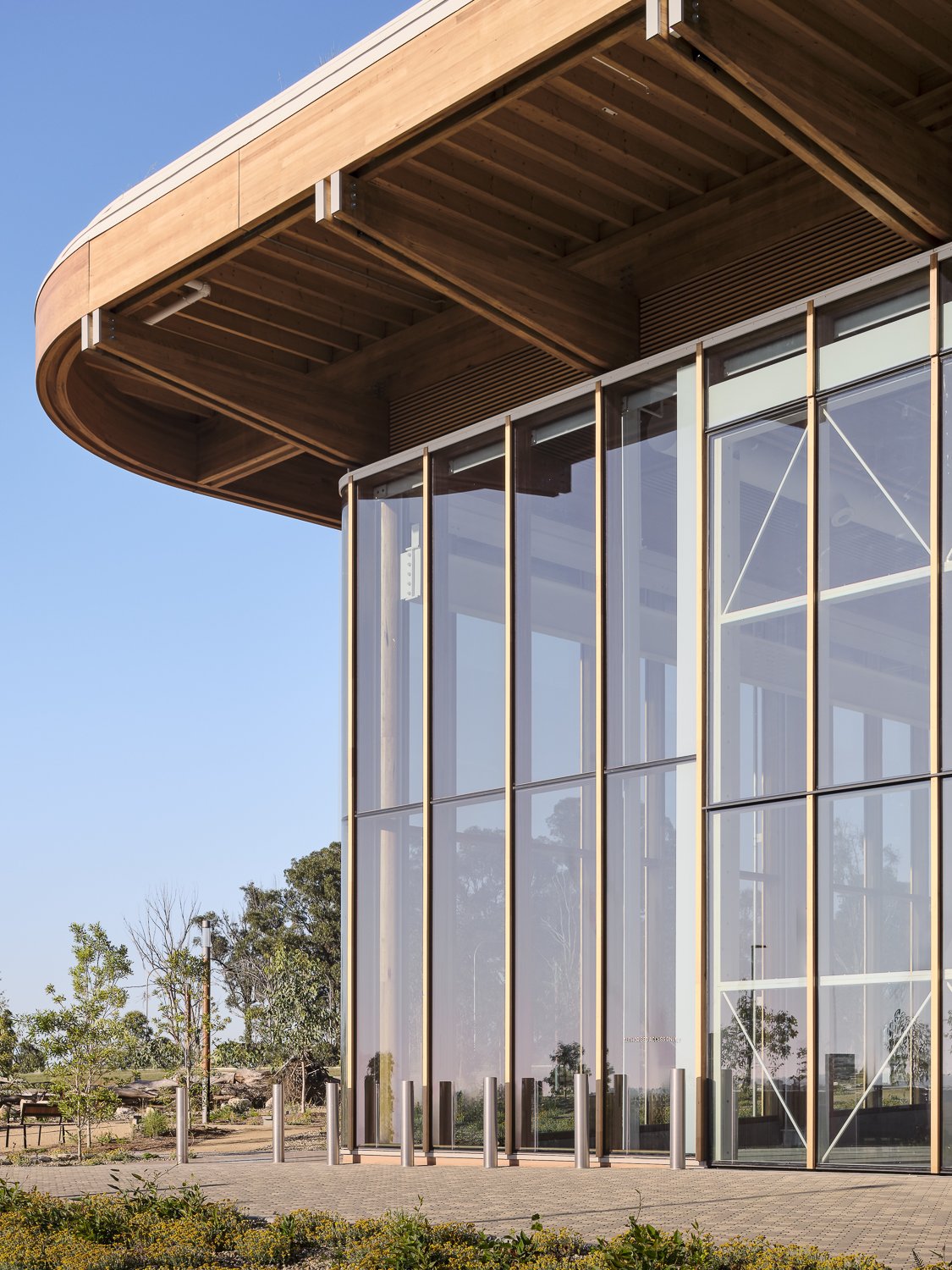 Modern building with large glass windows, wooden overhang, and outdoor landscaping under a clear blue sky.
