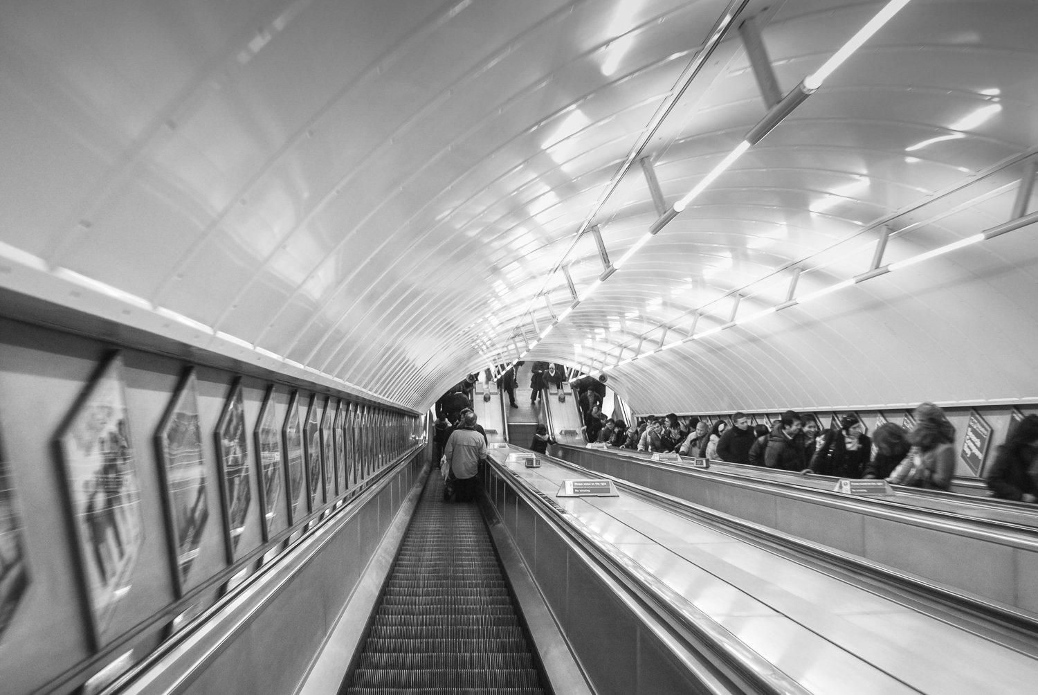 Black and white photo of a busy subway station with people on escalators and walking through the tunnel.