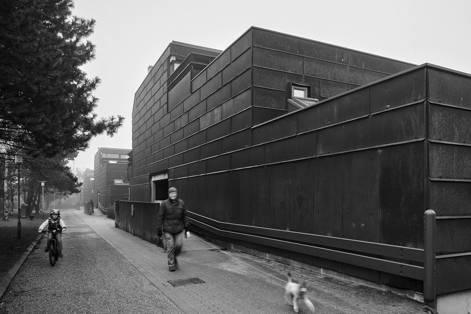 A black and white photo of a modern building with a person and a child riding a bike and walking a small dog on a sidewalk. There are trees and street lamps along the street.
