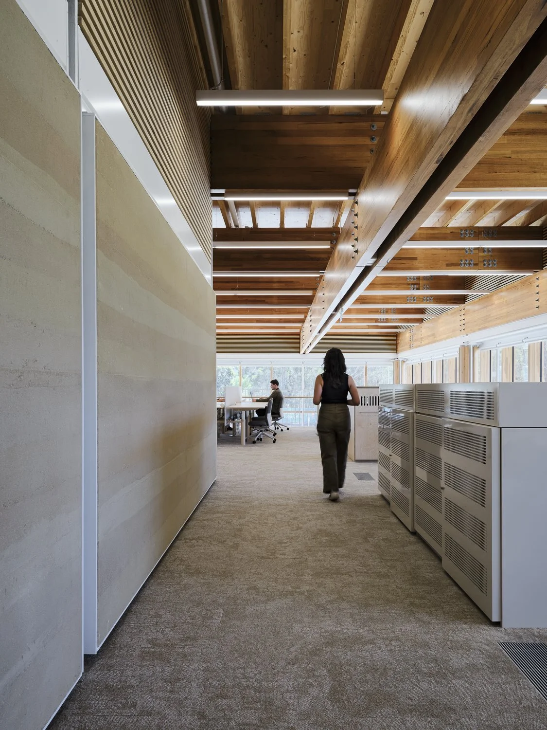 Interior of a modern office with wooden ceilings, large windows, and a woman walking down a hallway towards a workspace with a man working at his desk.
