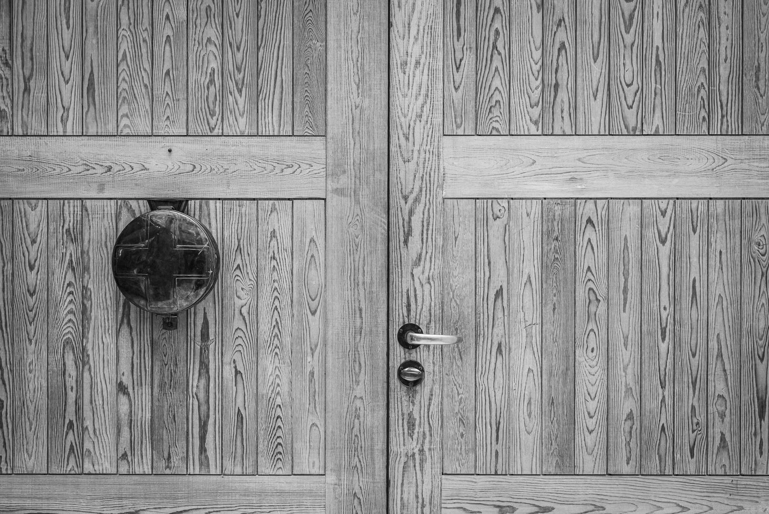 Black and white photo of wooden double doors with a door handle on the right door and a circular object attached to the left door.
